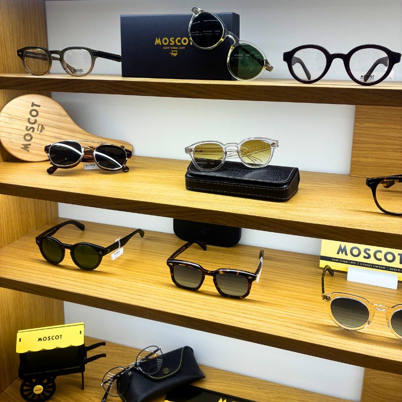 Présentation de lunettes de créateur Moscot sur des étagères en bois clair chez Bonot Opticiens. Exposition de montures optiques et solaires au design vintage et contemporain, avec des accessoires de la marque Moscot New York.